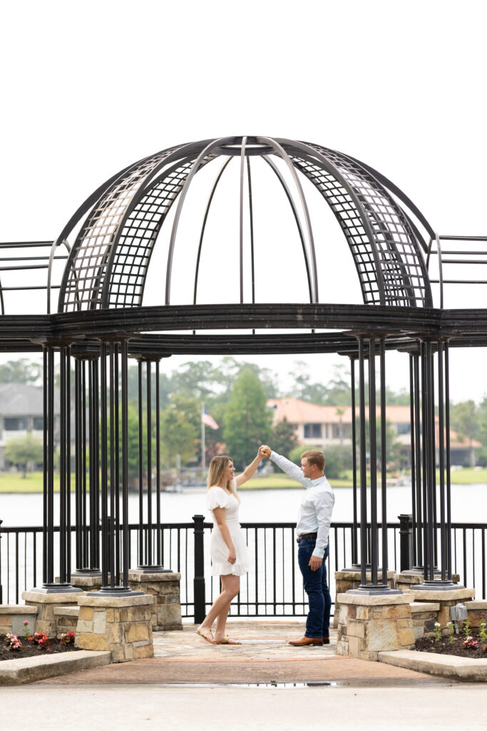 Couple dressed in jeans and white dress pretend to dance the woodlands engagement photos under a black iron structure